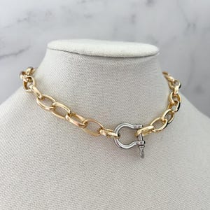 Puede incluir: Un collar de cadena dorada con un cierre plateado. El collar se exhibe sobre un busto de maniqu&iacute; blanco sobre un fondo de m&aacute;rmol. Los eslabones de la cadena son ovalados y gruesos, creando una pieza audaz.