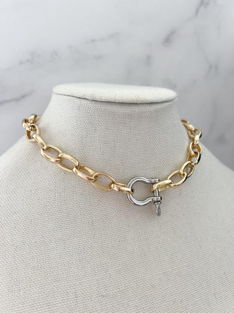 Puede incluir: Un collar de cadena dorada con un cierre plateado. El collar se exhibe sobre un busto de maniqu&iacute; blanco sobre un fondo de m&aacute;rmol. Los eslabones de la cadena son ovalados y gruesos, creando una pieza audaz.