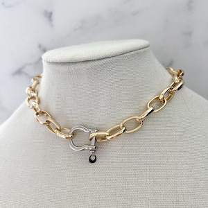 Puede incluir: Un collar de cadena dorada con un cierre plateado. El collar se exhibe sobre un busto de maniqu&iacute; blanco contra un fondo de m&aacute;rmol blanco y gris. Los eslabones de la cadena tienen forma ovalada.