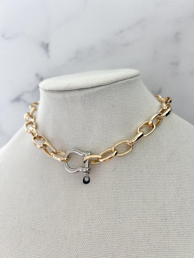 Puede incluir: Un collar de cadena dorada con un cierre plateado. El collar se exhibe sobre un busto de maniqu&iacute; blanco contra un fondo de m&aacute;rmol blanco y gris. Los eslabones de la cadena tienen forma ovalada.