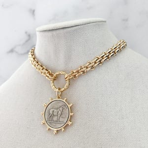 May include: A gold-toned chain necklace featuring a circular pendant with a silver-coloured horse relief. The pendant is framed by a sunburst-style border with small spheres. The chain consists of thick, interlocking links connected to a textured ring.