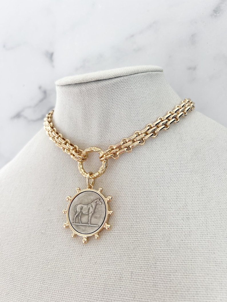 May include: A gold-toned chain necklace featuring a circular pendant with a silver-coloured horse relief. The pendant is framed by a sunburst-style border with small spheres. The chain consists of thick, interlocking links connected to a textured ring.