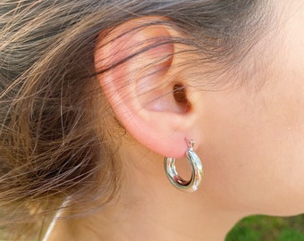 May include: Silver hoop earrings with a thick, rounded design. The earrings are worn in a person's ear.