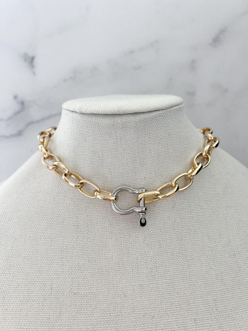 Puede incluir: Un collar de cadena dorada con un cierre plateado. El collar se exhibe en un maniqu&iacute;. Los eslabones de la cadena son ovalados y gruesos. El cierre tiene un dise&ntilde;o en forma de U con un peque&ntilde;o acento negro.