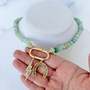 May include: A green jade beaded necklace with a gold clasp and charms. The charms include a horseshoe with green stones and a lightning bolt with clear stones.