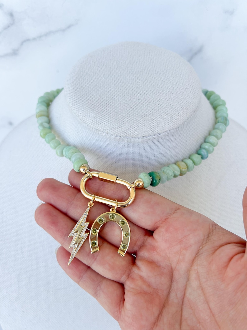 May include: A green jade beaded necklace with a gold clasp and charms. The charms include a horseshoe with green stones and a lightning bolt with clear stones.