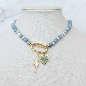 May include: A light blue gemstone beaded necklace with a gold clasp and two charms: a lightning bolt and a heart with blue crystals.