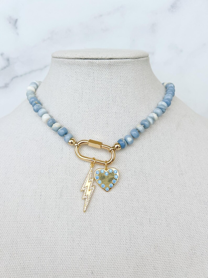 May include: A light blue gemstone beaded necklace with a gold clasp and two charms: a lightning bolt and a heart with blue crystals.