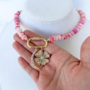 close up image of the gold carabiner featuring two gold charms - gold crescent moon necklace with pink stone detail and a gold four leaf clover pendant with clear stones