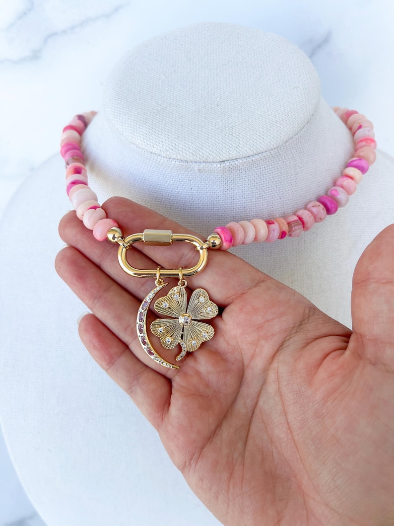 close up image of the gold carabiner featuring two gold charms - gold crescent moon necklace with pink stone detail and a gold four leaf clover pendant with clear stones