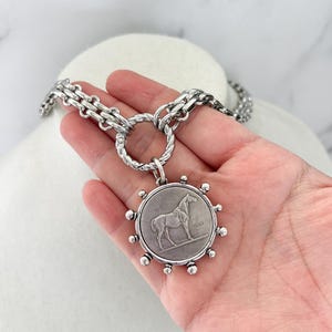May include: A silver-toned necklace featuring a thick, textured chain connected to a circular pendant. The pendant displays a relief of a standing horse and is surrounded by small, evenly spaced silver spheres along its outer edge.