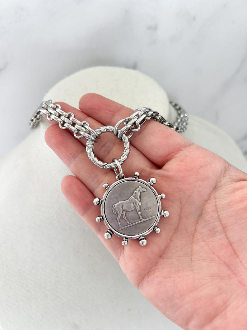 May include: A silver-toned necklace featuring a thick, textured chain connected to a circular pendant. The pendant displays a relief of a standing horse and is surrounded by small, evenly spaced silver spheres along its outer edge.