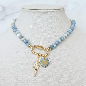 Blue opal beaded necklace with a large gold carabiner clasp in the front a gold lighting bolt charm with clear stone details and a gold heart charm with blue stones is attached to the carabiner