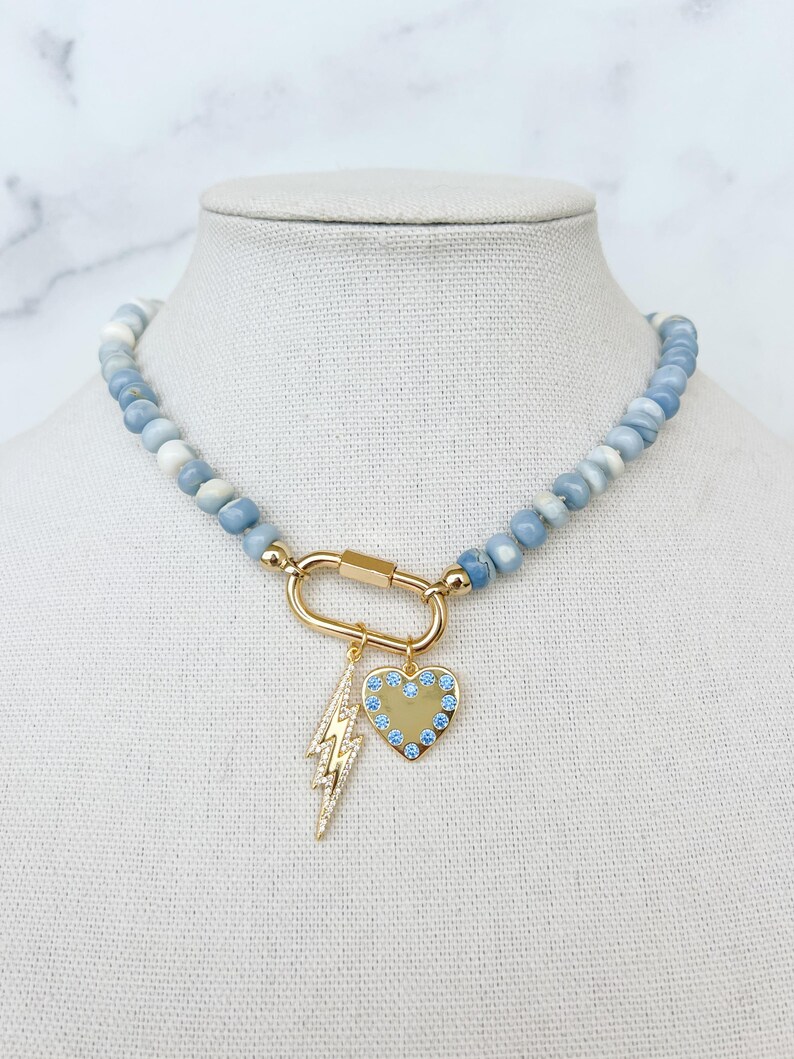 Blue opal beaded necklace with a large gold carabiner clasp in the front a gold lighting bolt charm with clear stone details and a gold heart charm with blue stones is attached to the carabiner