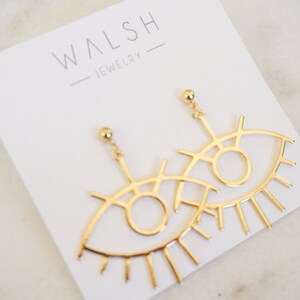 May include: Gold eye-shaped earrings with a delicate, intricate design. The earrings feature a large, open circle representing the eye, surrounded by smaller lines and curves. The bottom of the earrings have a fringe of thin, pointed lines resembling eyelashes.