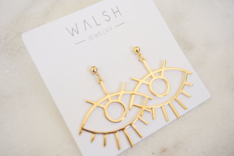 May include: Gold eye-shaped earrings with a delicate, intricate design. The earrings feature a large, open circle representing the eye, surrounded by smaller lines and curves. The bottom of the earrings have a fringe of thin, pointed lines resembling eyelashes.