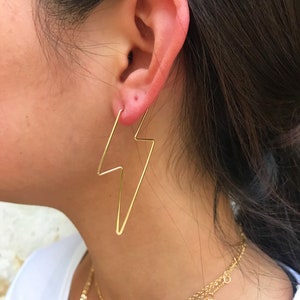 May include: Gold lightning bolt hoop earrings. The earrings are worn in the left ear of a person with long dark hair.