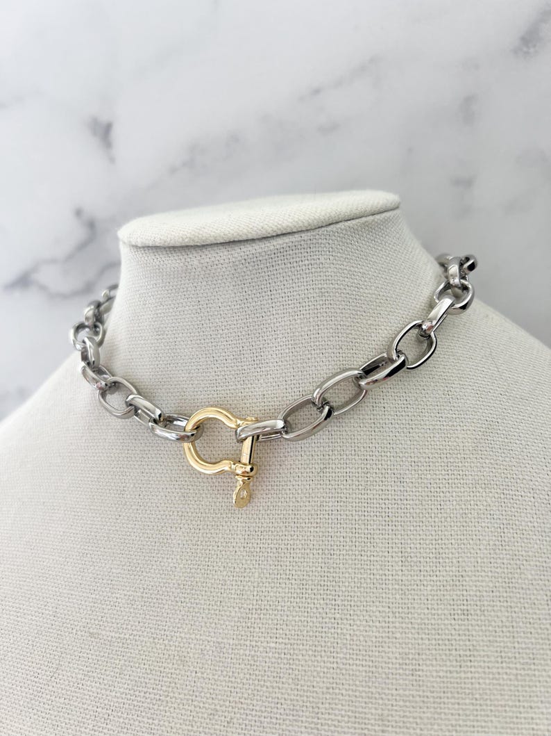 Puede incluir: Collar de cadena plateada con cierre dorado. El collar se exhibe sobre un busto de maniqu&iacute; blanco sobre un fondo de m&aacute;rmol. Los eslabones de la cadena son de forma ovalada.