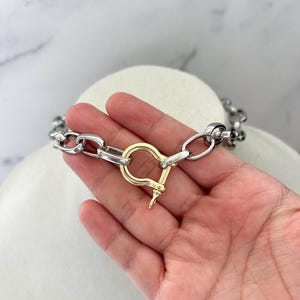 Puede incluir: Un collar de cadena plateada con un cierre dorado. El collar se exhibe sobre una superficie blanca. El cierre tiene un dise&ntilde;o &uacute;nico, agregando un toque de elegancia a la joya.