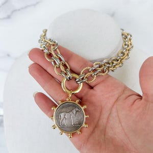 May include: A gold and silver chain necklace with a large round pendant featuring a horse design. The pendant has a textured silver surface and a gold rim with small gold balls around the edge.