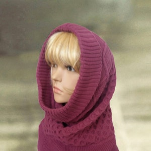 May include: A close-up of a pink knitted cowl neck with a hood. The cowl neck is folded over and has a cable knit pattern. The hood is pulled up over the head of a mannequin.