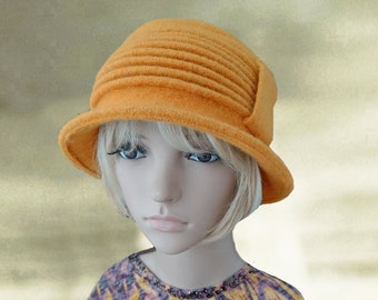 May include: An orange cloche hat with a rounded crown and a small brim. The hat has a textured, pleated design and is made of a soft material. The hat is displayed on a mannequin with blonde hair and a patterned top.