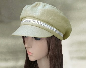May include: A light green linen newsboy cap with a white ribbon trim featuring a repeating text pattern. The cap has a slightly curved brim and a rounded crown.