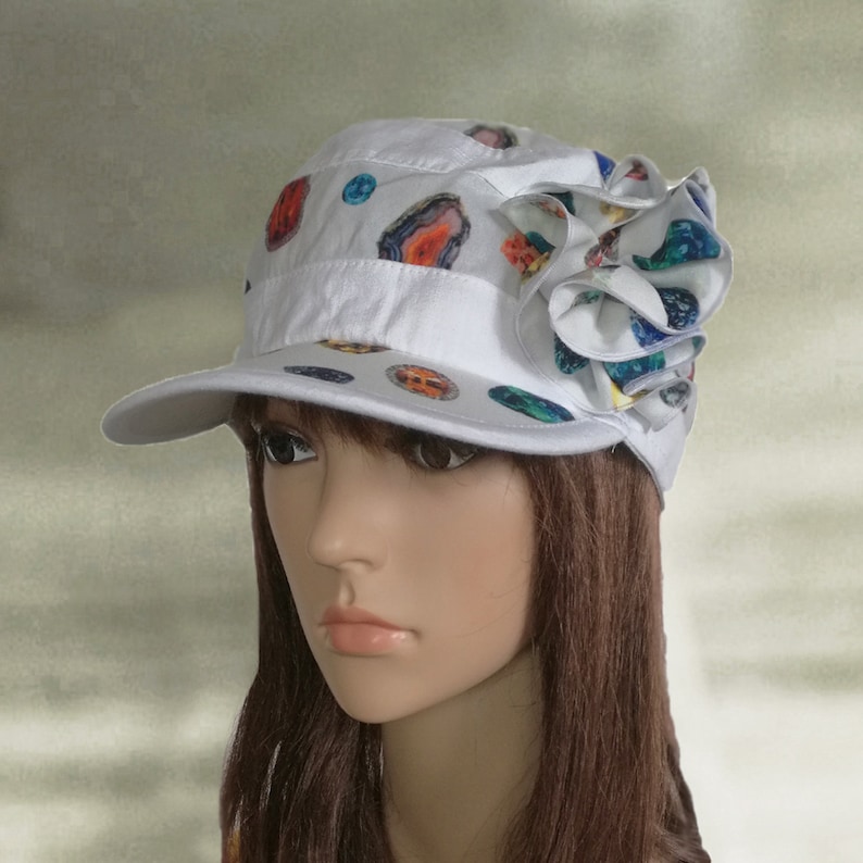 May include: White cotton cap with a colorful flower design. The cap has a short brim and a patterned design of colorful circles and swirls.