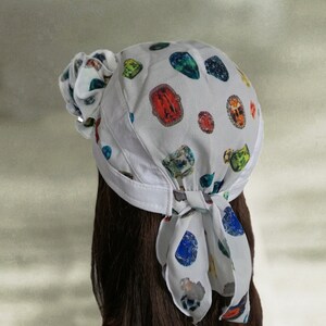May include: White bandana with a colorful gemstone print. The bandana is tied at the back of the head.
