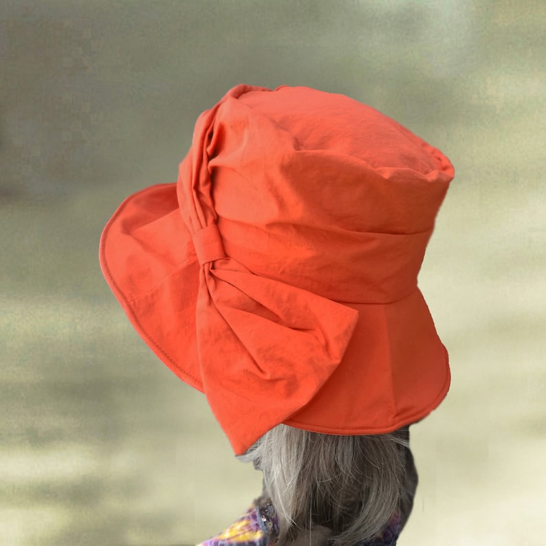 Puede incluir: Un sombrero de sol naranja brillante con un gran lazo en un lado. El sombrero est&aacute; hecho de una tela ligera y tiene un ala ancha.