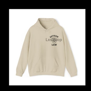 Op de afbeelding: Een beige hoodie met een zwarte grafische print van een schild met een weegschaal van de rechtvaardigheid en de tekst "Gotham Law".