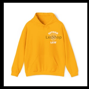 Op de afbeelding: Een gele hoodie met een witte afbeelding op de voorkant met de tekst "Gotham Law" met een schild en kroonontwerp.