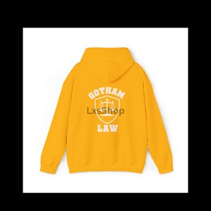 Op de afbeelding: Een gele hoodie met een witte afbeelding op de rug die "Gotham Law" zegt met een weegschaal van de rechtvaardigheid in een schild.