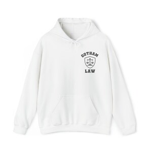 Op de afbeelding: Witte hoodie met een zwarte en grijze afbeelding van een schild met een weegschaal en de tekst "Gotham Law" op de voorkant.