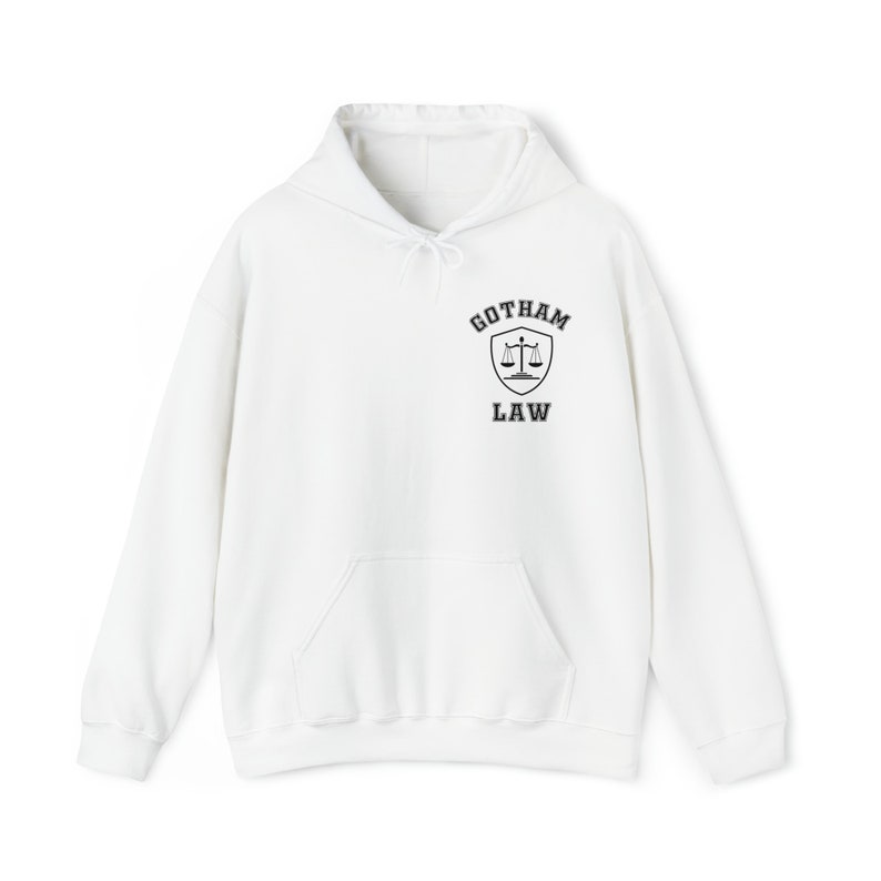 Op de afbeelding: Witte hoodie met een zwarte en grijze afbeelding van een schild met een weegschaal en de tekst "Gotham Law" op de voorkant.