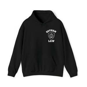 Op de afbeelding: Zwart hoodie met een witte afbeelding van een schild met een weegschaal van de rechtvaardigheid en de tekst "Gotham Law" erop.