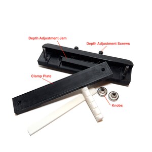 K&ouml;nnte beinhalten: Eine schwarze, im 3D-Druck hergestellte Gitarren-Steg-Klemme mit einem wei&szlig;en Sattel aus Kunststoff. Die Klemme besteht aus zwei schwarzen Platten, eine mit einer Tiefenverstell-Sperre und die andere mit Tiefenverstellschrauben. Die Klemme enth&auml;lt au&szlig;erdem zwei silberne Kn&ouml;pfe.