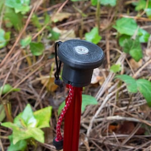 May include: A close-up of a black trekking pole tip with a silver screw. The pole is red and has a black cord attached to it.