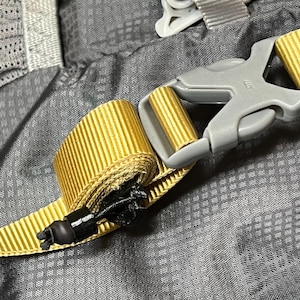 May include: Close-up of a yellow and gray adjustable strap with a black buckle. The strap is attached to a gray fabric background.