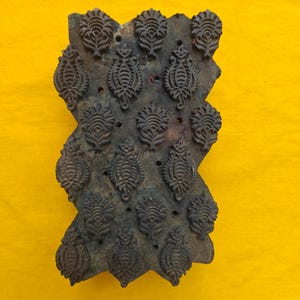 May include: A rectangular dark brown wooden printing block featuring a repeating carved pattern of stylised floral motifs. The surface displays a weathered texture with small circular indentations interspersed between the raised carved designs.