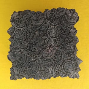 May include: A dark grey, intricately carved wooden block with a floral and leaf pattern. The design features detailed flowers and foliage, ideal for textile printing or decorative purposes. The block is square-shaped.