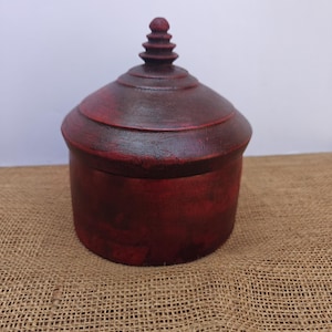Può includere: Una scatola rotonda in legno rosso scuro con coperchio abbinato. Il coperchio ha una forma arrotondata con un elemento decorativo sulla parte superiore. La scatola è appoggiata su una superficie di iuta. La scatola viene probabilmente utilizzata per la conservazione.