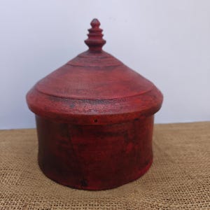 Può includere: Una scatola di legno rossa fatta a mano con coperchio a cupola e un terminale decorativo. La scatola ha una base cilindrica e una superficie leggermente strutturata. Il design generale è semplice ed elegante, adatto per riporre piccoli oggetti. La scatola è alta circa 15 cm.