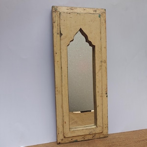 May include: A rectangular wall mirror featuring a distressed, cream-colored wooden frame with a pointed arch cutout at the top. The frame displays visible signs of wear, including chipped paint and surface imperfections, surrounding a vertical glass pane.