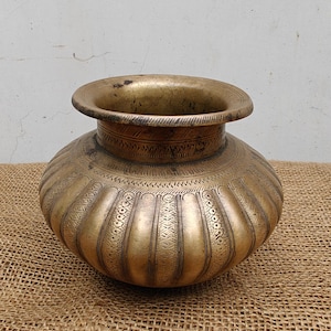 May include: A vintage brass pot with a wide, flared rim and a rounded body. The pot features vertical ribbed detailing and intricate etched patterns around the neck and body. The aged brass has a warm, golden hue.