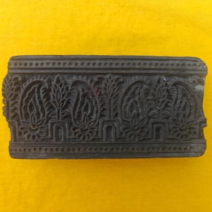 Puede incluir: Un bloque de impresión rectangular de madera marrón oscuro con un patrón tallado repetitivo de motivos de cachemira y elementos florales estilizados, bordeado por líneas horizontales con pequeñas muescas circulares.