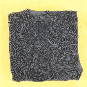May include: A dark grey, rectangular wooden block with an intricate floral and paisley design carved into its surface. The block features detailed patterns of flowers, leaves, and swirling lines, ideal for textile printing or decorative crafts.