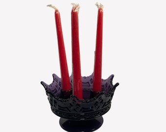 May include: A dark purple glass candle holder with a crown design, holding three red taper candles. The candle holder has a decorative pattern around the rim and sits on a round base. The candles are unwaxed.