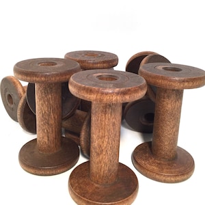 May include: A collection of vintage wooden spools, each with a circular base and top connected by a cylindrical stem. The spools are a warm brown color, showing the natural wood grain. Some spools are stacked, others are standing.