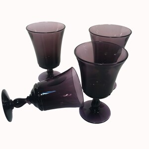 May include: A set of three purple glass goblets with a smooth, translucent finish. Two of the goblets are standing upright on circular bases, while the third goblet is positioned horizontally on its side, showcasing the stem and bowl design.
